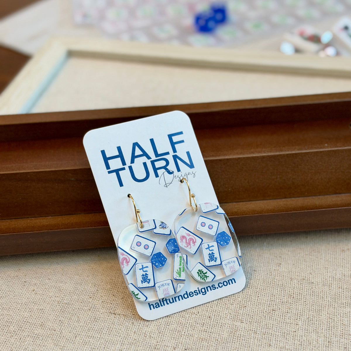 Blue chinoiserie mahjong tile earrings in clear acrylic with Jokers and dice on gold-tone hooks, beside mahjong tiles and racks