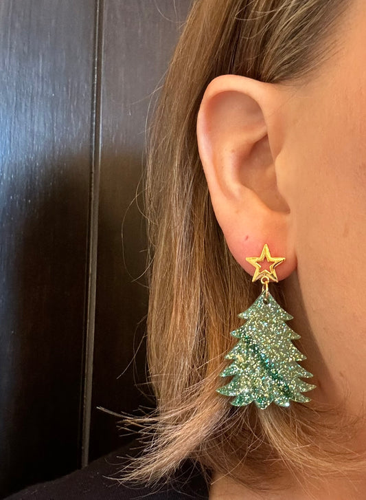 Model wearing Green and gold glitter acrylic Christmas tree earrings with gold star studs hanging on display rod, showing sparkling festive dangle design by Half Turn Designs