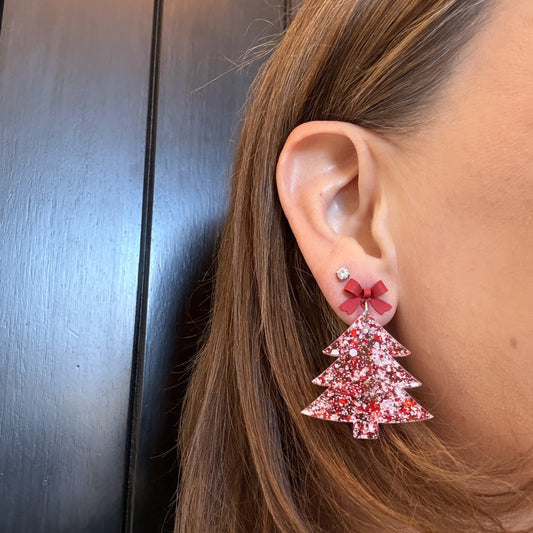 Model wearing glittery Red and white acrylic Christmas tree earrings with red bow studs, lightweight with 2 inch drop length, holiday dangles styled on a neutral background.