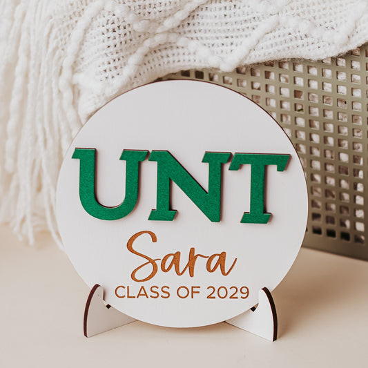 Officially licensed University of North Texas 5 inch round wooden dorm decor, shelf sitter and desk sign in white wood with the UNT letters in Mean Green, personalized with your choice of 1-2 lines of engraved text in four wood finishes. Comes with a matching stand.
