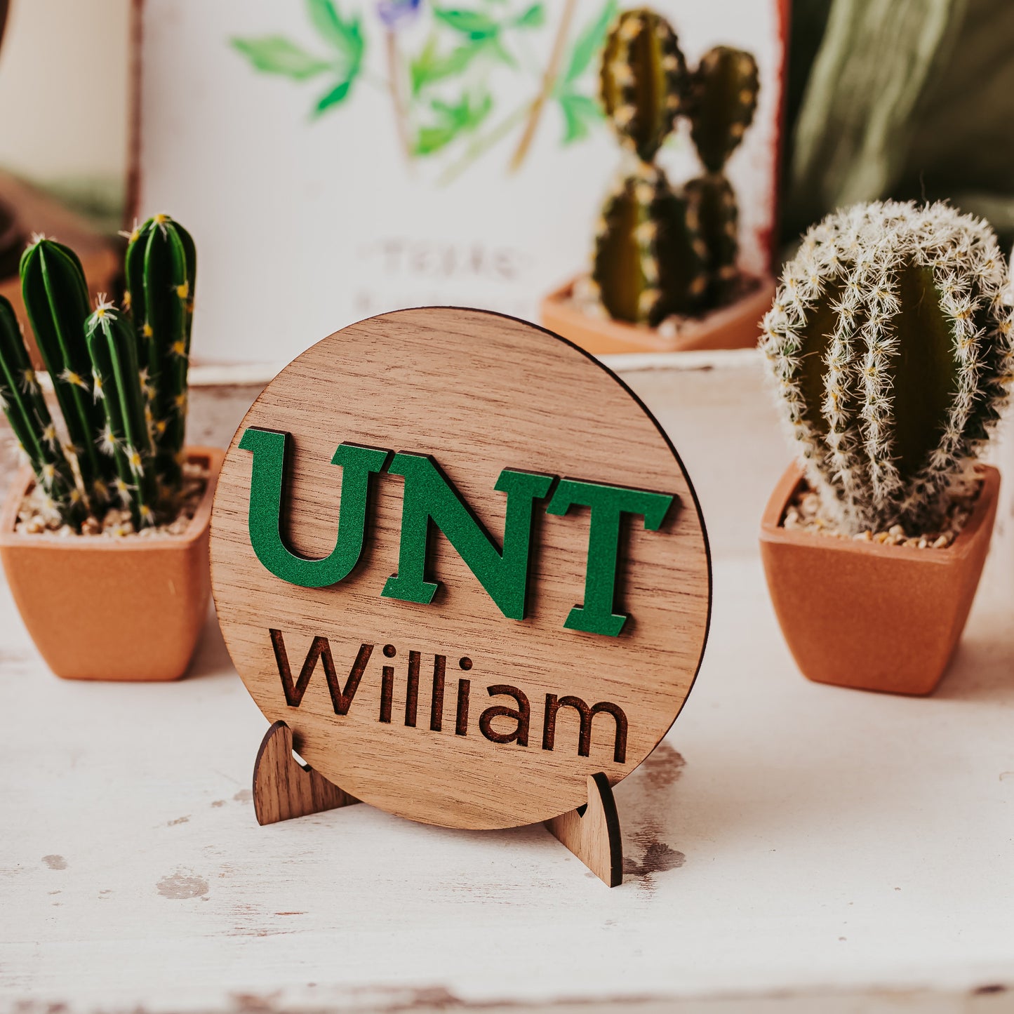 Officially licensed University of North Texas 5 inch round wooden dorm decor, shelf sitter and desk sign in walnut wood finish with the UNT letters in Mean Green, personalized with your choice of 1-2 lines of engraved text in four wood finishes. Comes with a matching stand.