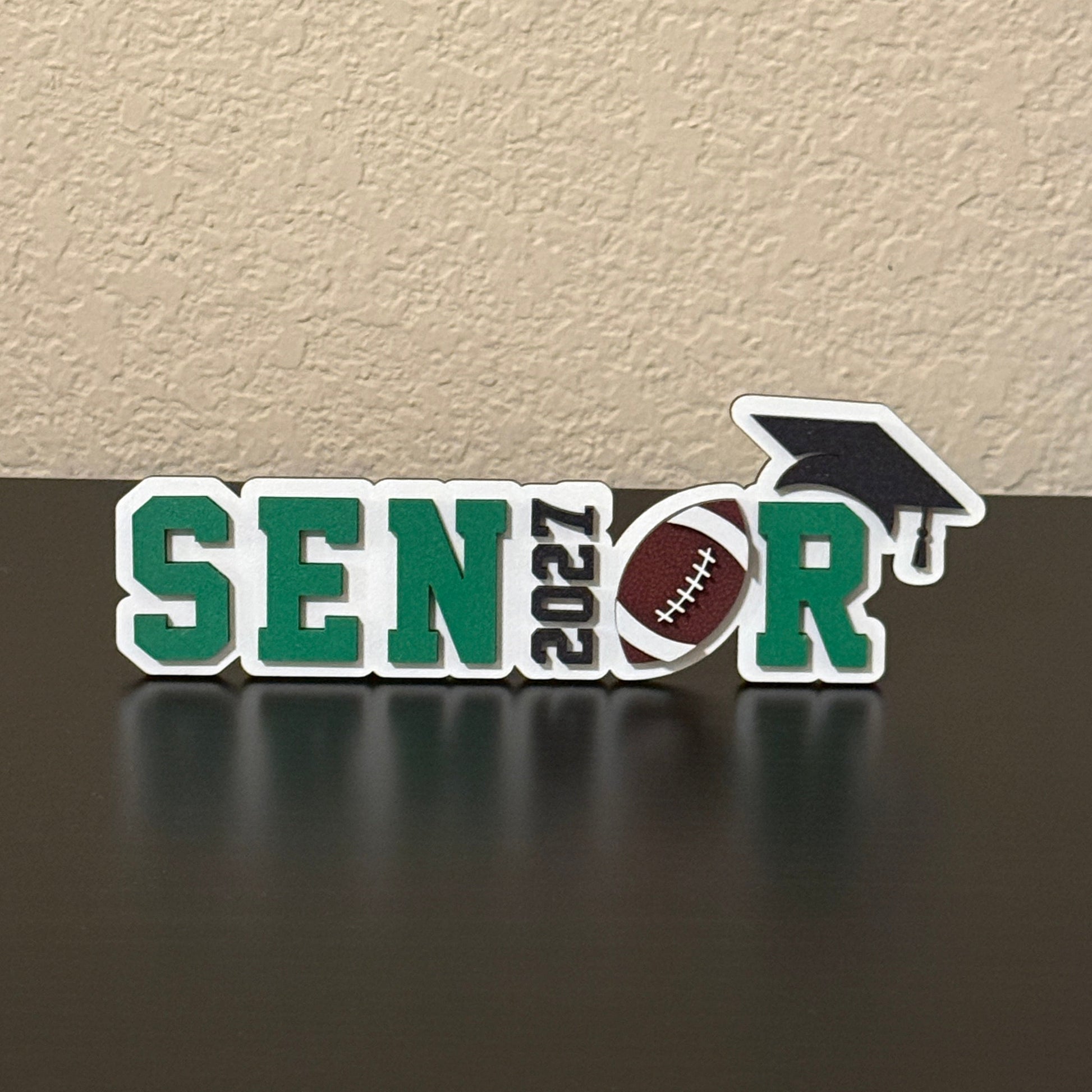 Class of 2027 football senior sign with white wood base, green letters, 2027 as the I, football as the O, and black cap.