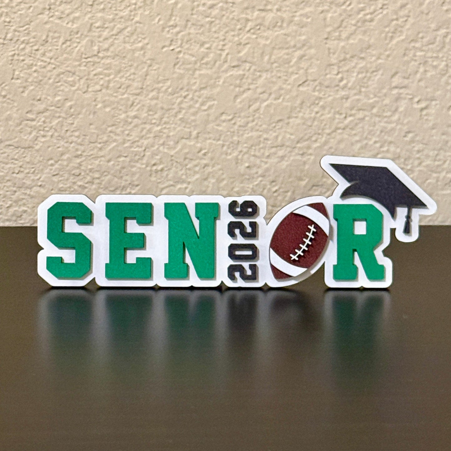 Class of 2026 High School Senior football sign measuring 7" in length by 2.35 inches in height.   White wood background with black graduation cap and 2026 letters, brown and white football and customers choice of colors for the letters SENR, which is part of the wood senior, perfect for graduation parties and football banquets.
