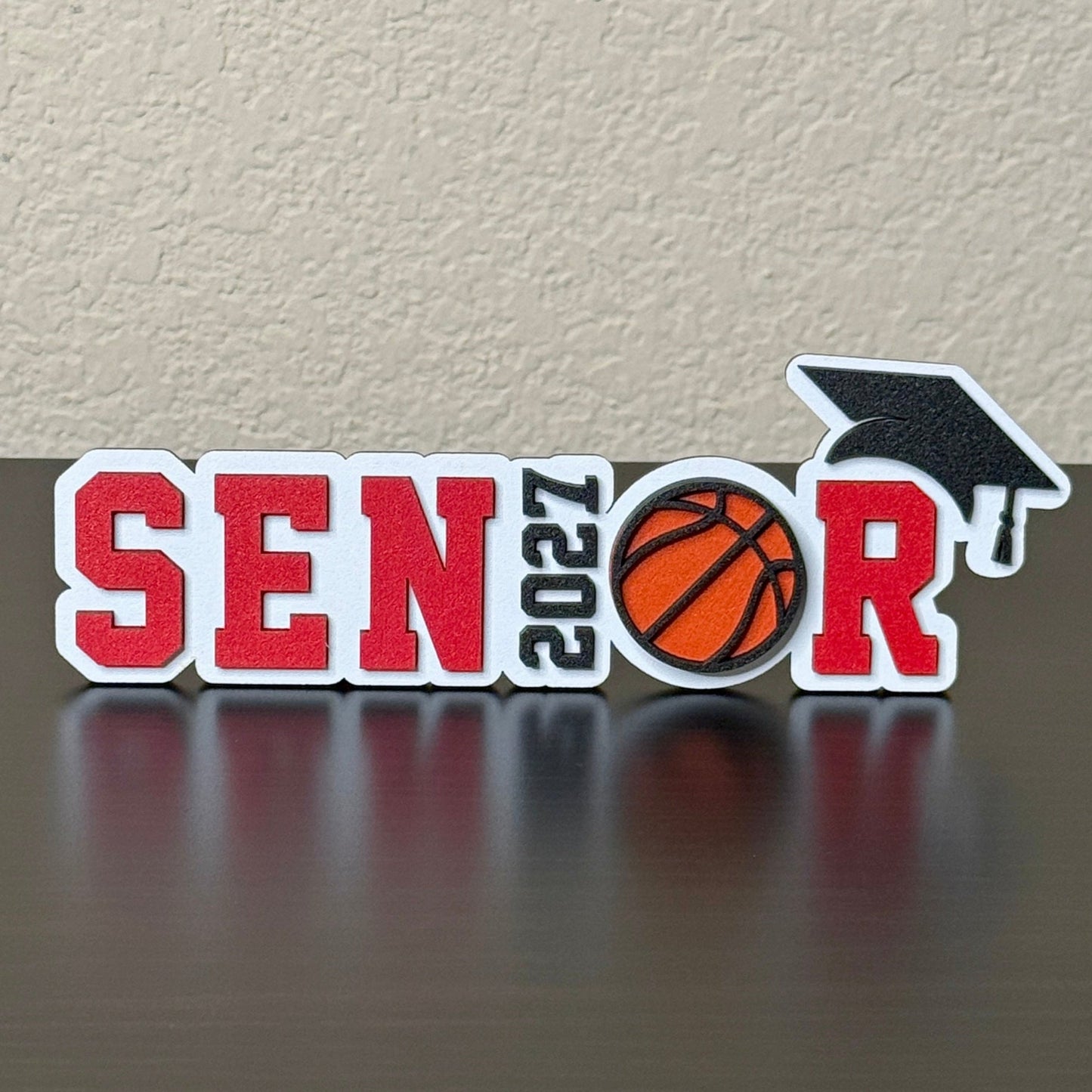 Class of 2027 basketball senior sign with red letters, orange basketball, black graduation cap, and white wood base.