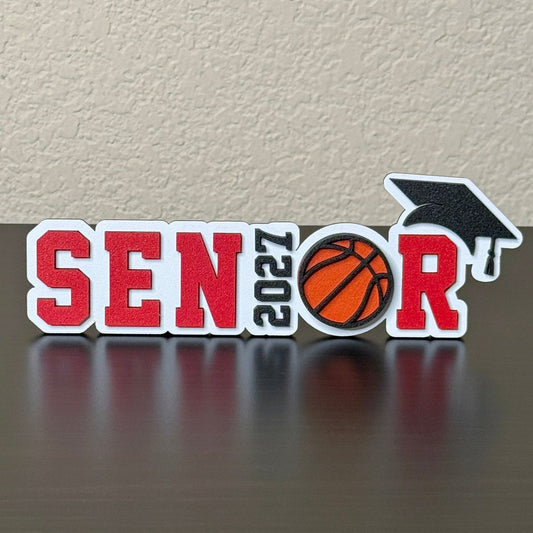 Class of 2027 basketball senior sign with red letters, orange basketball, black graduation cap, and white wood base.