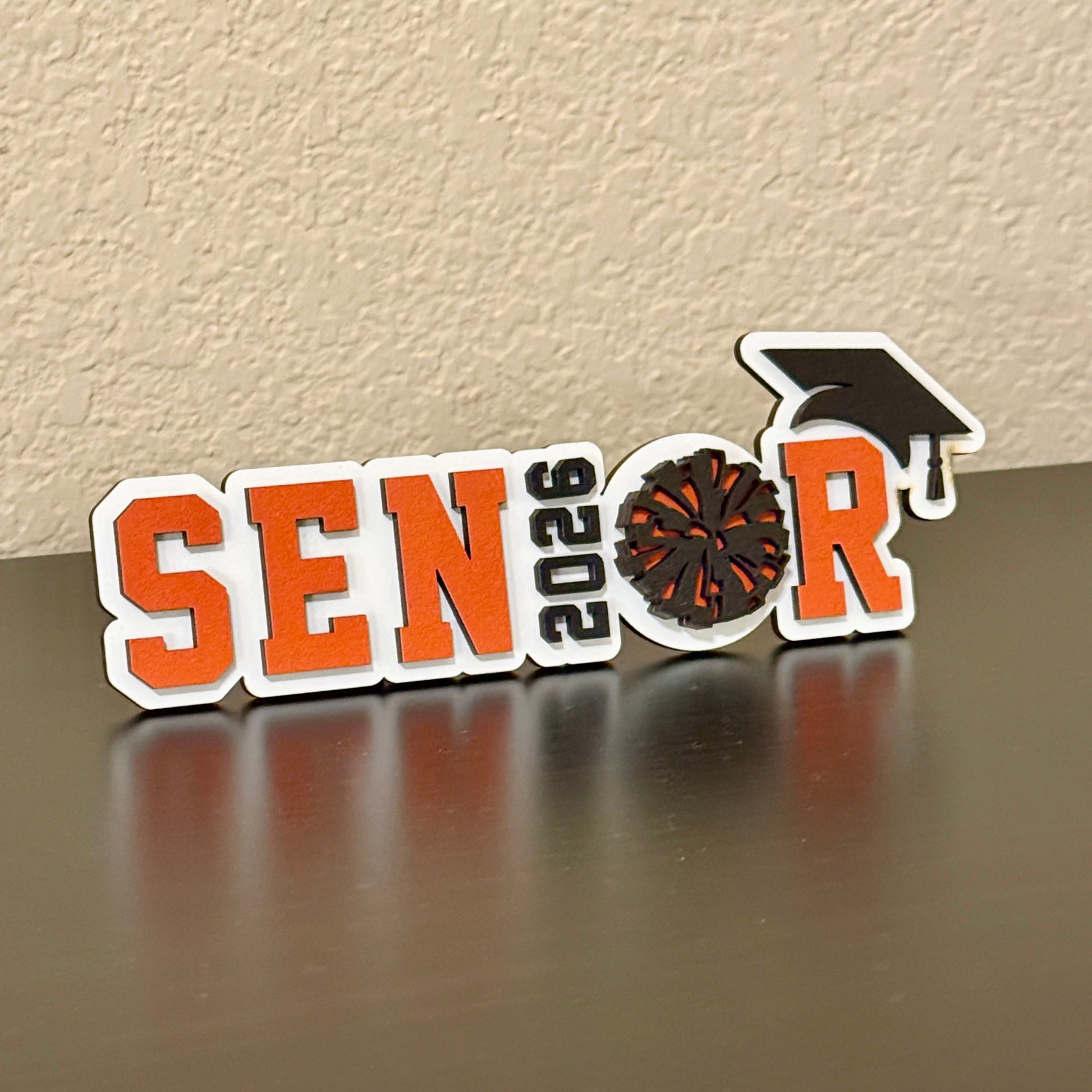 Class of 2026 High School Senior Graduation Sign for Pom and Drill Team members in white wood with orange and black accents.  Spells SENIOR with 2026 as the "I" and a two-layer pom pom as the "O" with a graduation hat.