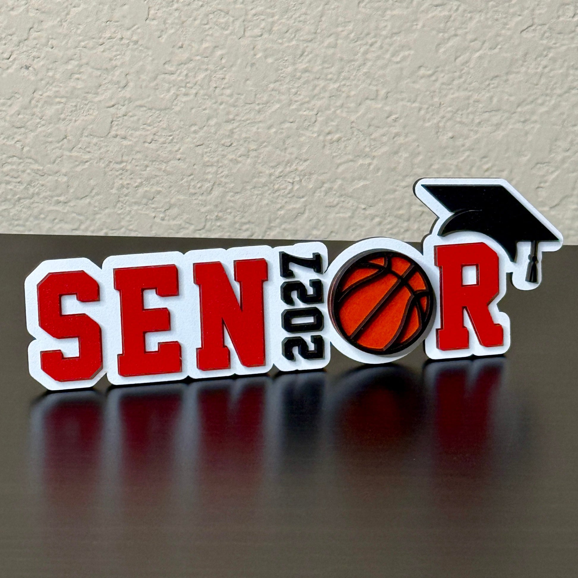 Class of 2027 basketball senior sign with red letters, orange basketball, black graduation cap, and white wood base.