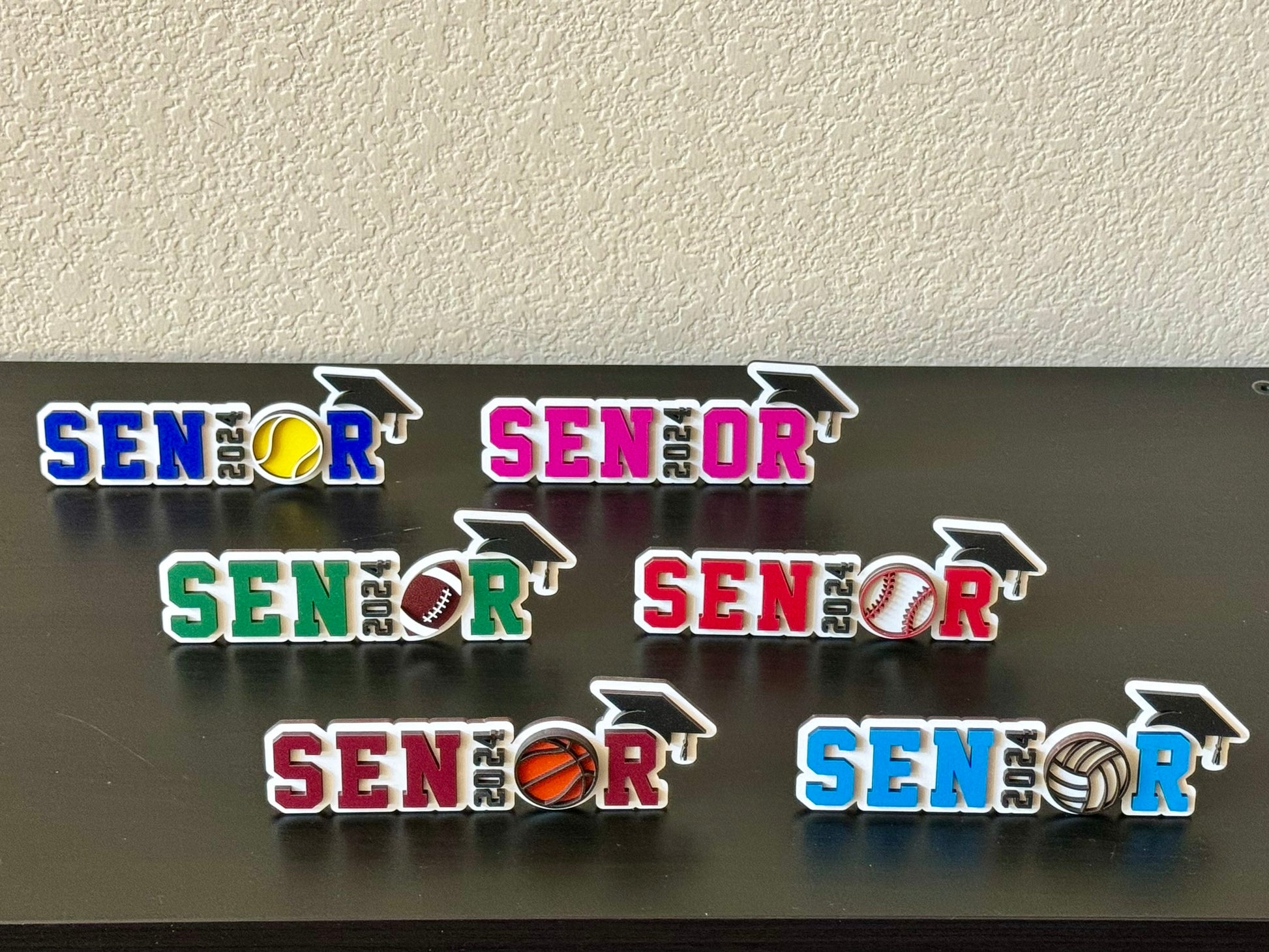 High School Senior spots signs measuring 7" in length by 2.35 inches in height.   White wood background with black graduation cap and year of graduation numbers, sports symbol o sport appropriate colors and customers choice of colors for the letters SENR, which is part of the wood senior, perfect for graduation parties and sports banquets.