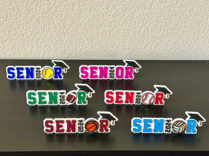 High School Senior spots signs measuring 7" in length by 2.35 inches in height.   White wood background with black graduation cap and year of graduation numbers, sports symbol o sport appropriate colors and customers choice of colors for the letters SENR, which is part of the wood senior, perfect for graduation parties and sports banquets.