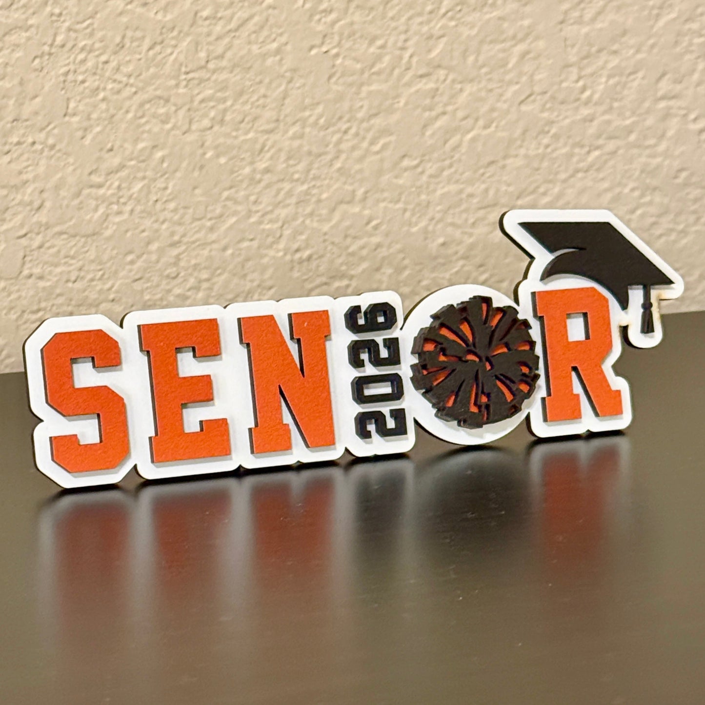 High School Cheerleader Graduation sign with white wood base. Spells out Senior with the graduation year as the "I" and the Pom Pom as the "O", with a black graduation cap.  Senior letters available in one of 14 school colors