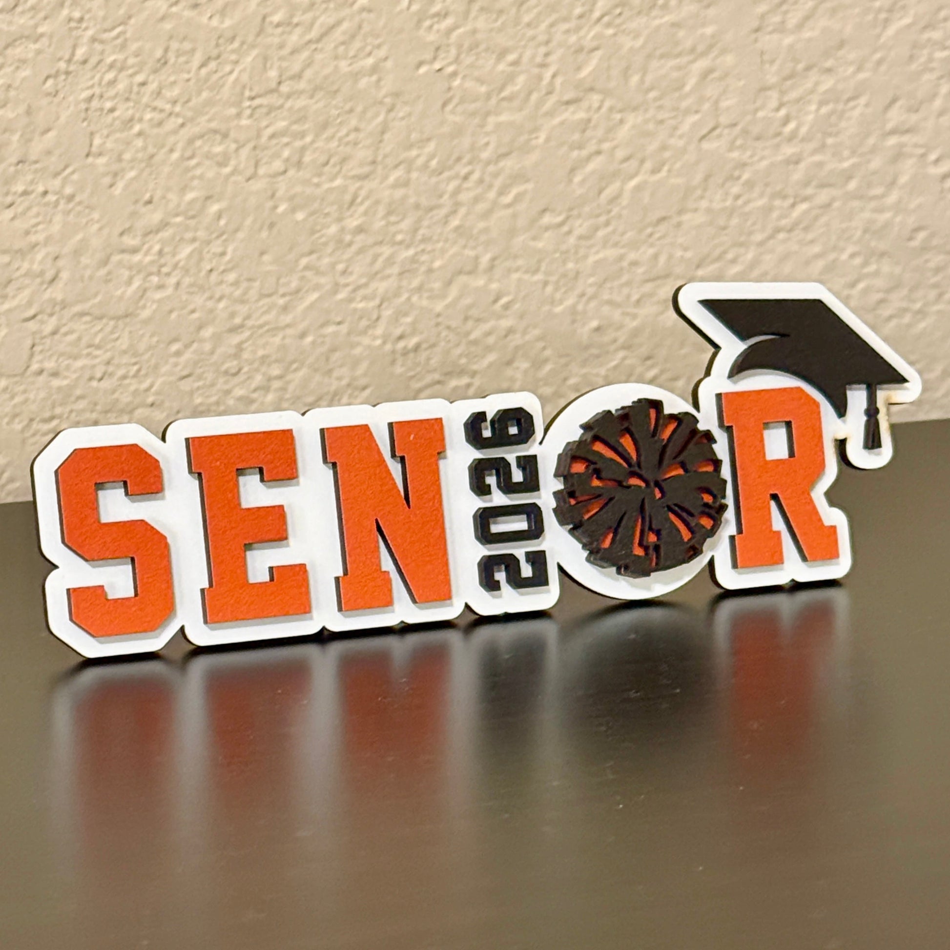 High School Cheerleader Graduation sign with white wood base. Spells out Senior with the graduation year as the "I" and the Pom Pom as the "O", with a black graduation cap.  Senior letters available in one of 14 school colors