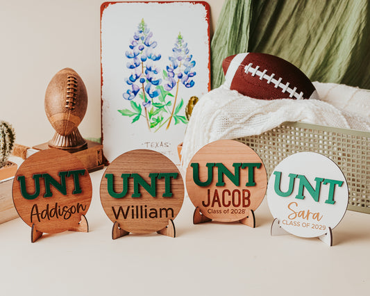 Officially licensed University of North Texas 5 inch round wooden dorm decor, shelf sitter and desk sign with the UNT letters in Mean Green, personalized with your choice of 1-2 lines of engraved text in four wood finishes. From left to right the finishes shown here are mahogany, walnut, cherry and white.  Comes with a matching stand.