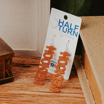 A pair of University of Texas Longhorns glitter dangle earrings on Half Turn Designs display card, featuring burnt orange and white acrylic word designs that spell “HOOK ’EM” on silver hooks