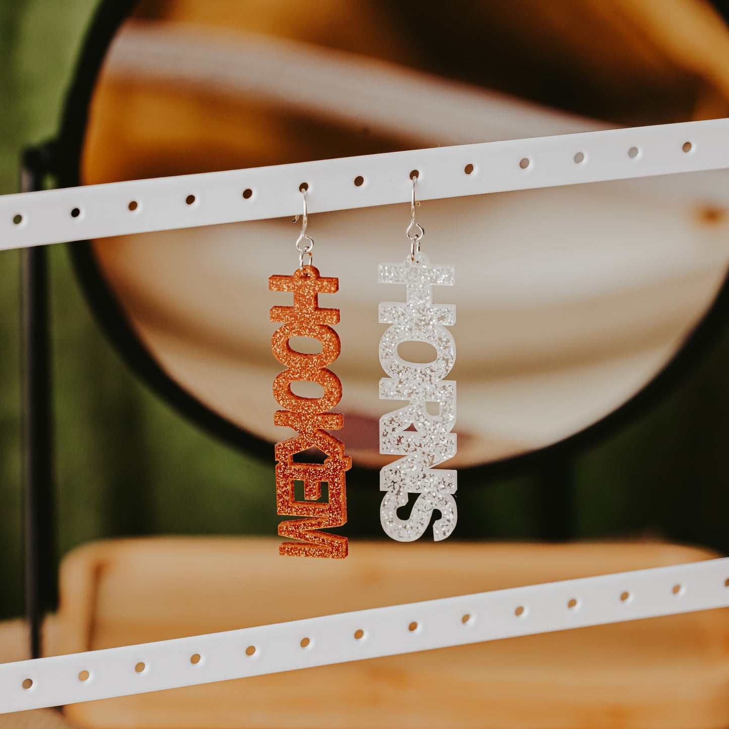 A pair of University of Texas Longhorns glitter dangle earrings on Half Turn Designs display card, featuring burnt orange and white acrylic word designs that spell “HOOK ’EM” and "HORNS"