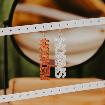 A pair of University of Texas Longhorns glitter dangle earrings on Half Turn Designs display card, featuring burnt orange and white acrylic word designs that spell “HOOK ’EM” and "HORNS"