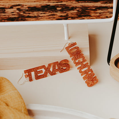 A pair of University of Texas Longhorns glitter dangle earrings  featuring burnt orange acrylic word designs that spell “TEXAS” and “LONGHORNS” on silver hooks