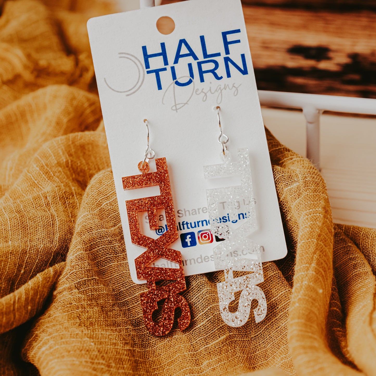 A pair of University of Texas Longhorns glitter dangle earrings on Half Turn Designs display card, featuring burnt orange and white acrylic word designs that spell “TEXAS” on silver hooks