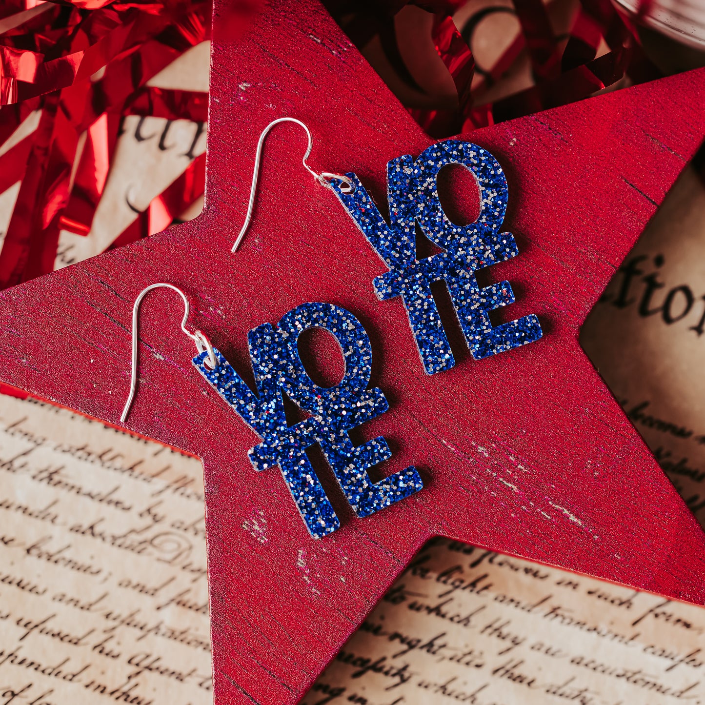 Blue glitter vote earrings with stacked VOTE letters and silver ear wires displayed on a red wooden star