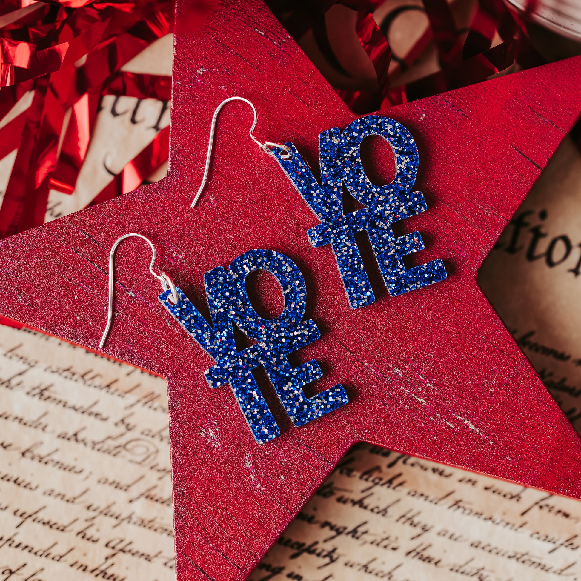 Blue glitter vote earrings with stacked VOTE letters and silver ear wires displayed on a red wooden star