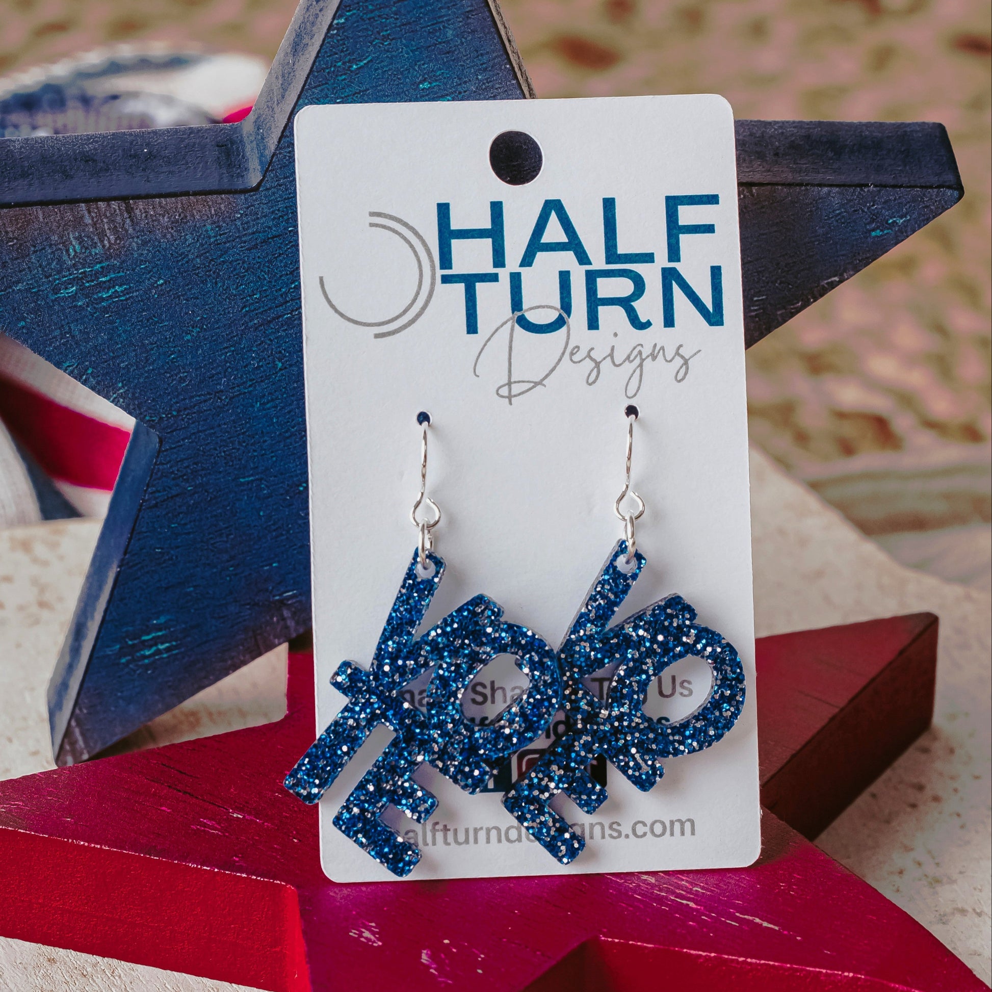 Blue glitter vote earrings with stacked VOTE letters and silver ear wires displayed on a red wooden star