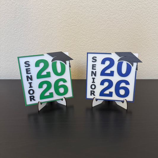Wooden Class of 2026 senior graduation signs in white with blue and green school color accents and matching stands displayed on a table