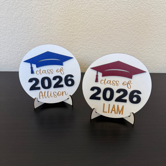 Personalized Class of 2026 senior signs with custom name on white wood round sign with stand. Shown here with blue and maroon accents