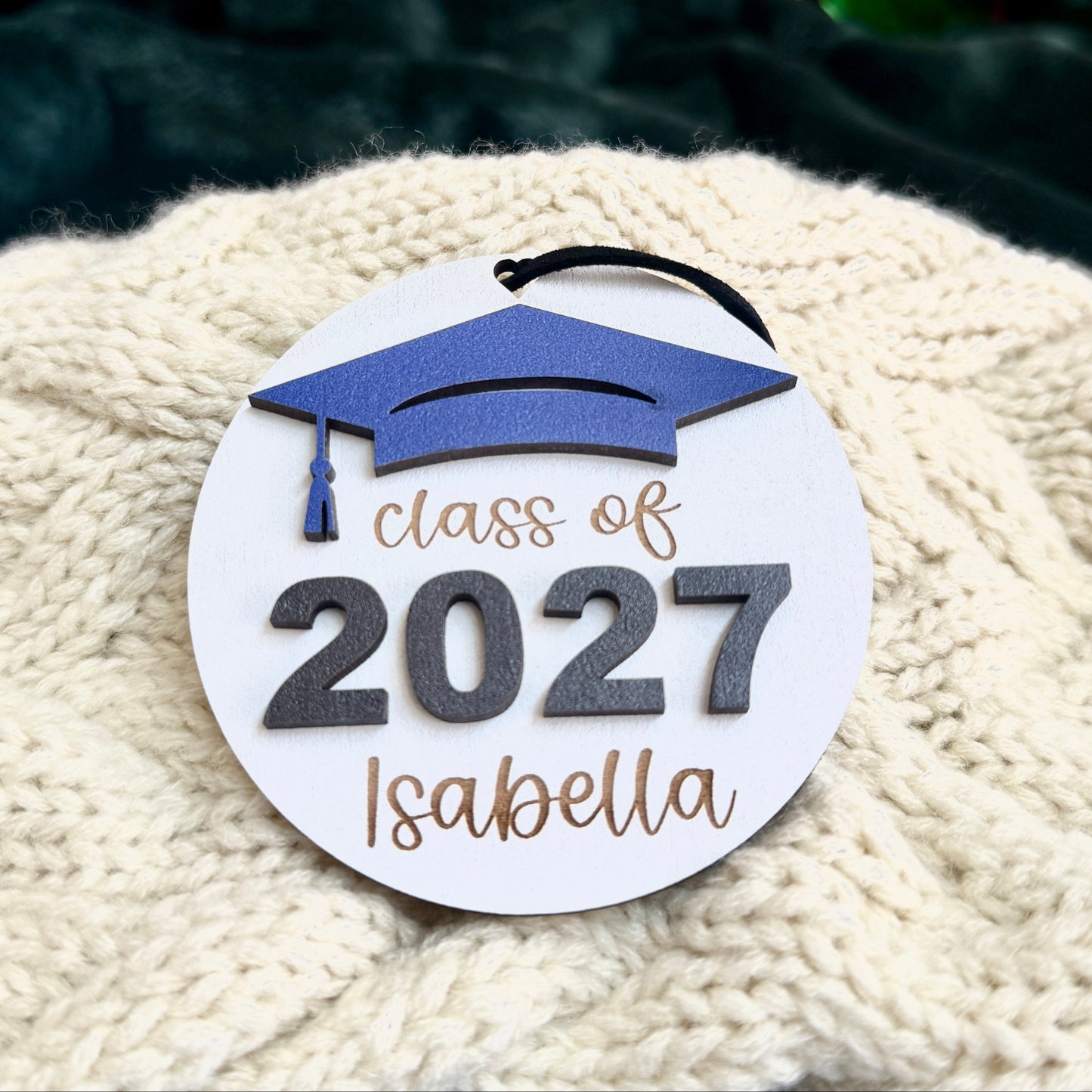 Personalized Class of 2027 wooden ornament with blue graduation cap, engraved graduate's name, raised 2027 numbers with a black cord hanger on white fabric