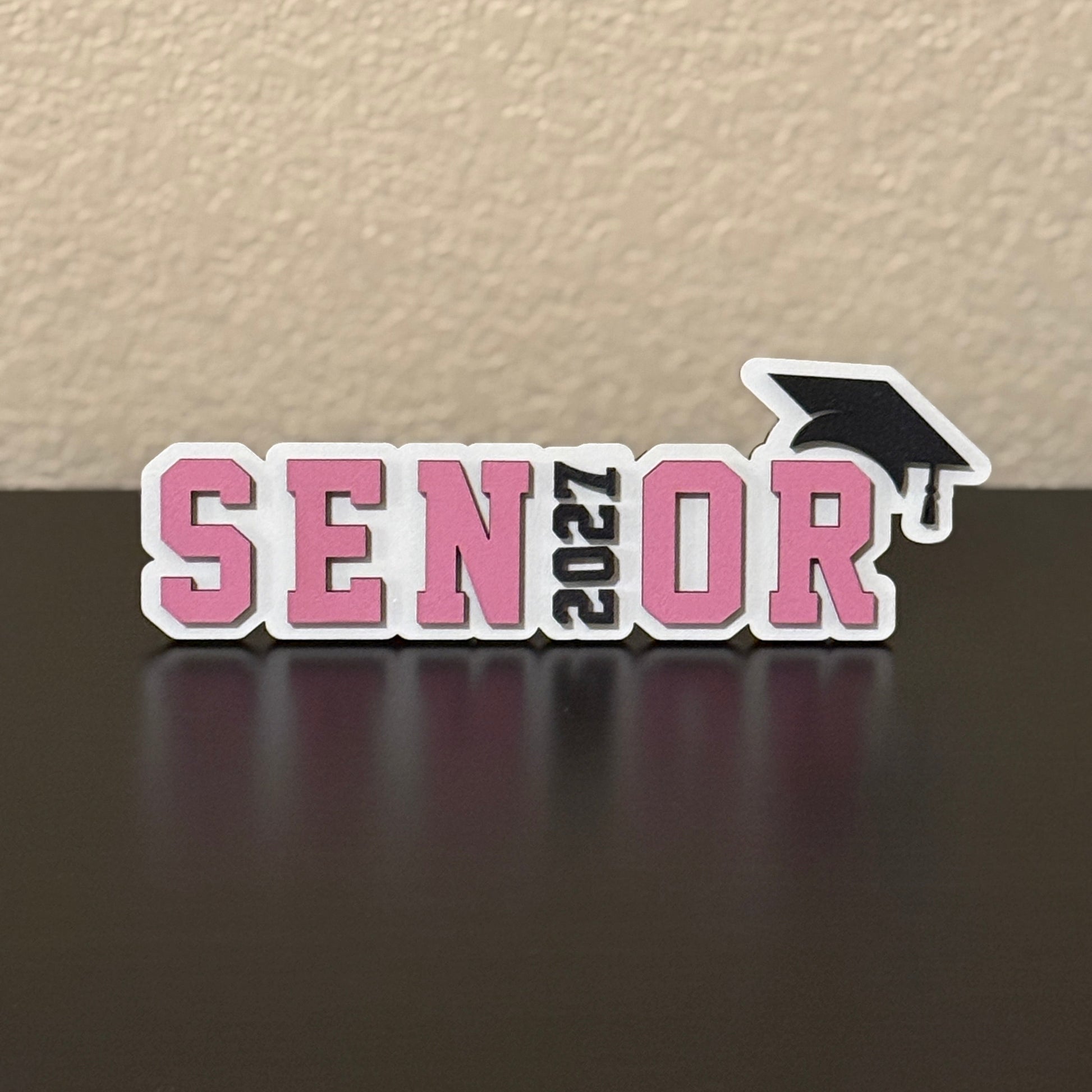Class of 2027 white wood senior sign standing on a tabletop, with pink SENIOR letters, 2027 replacing the I with a black graduation cap detail.