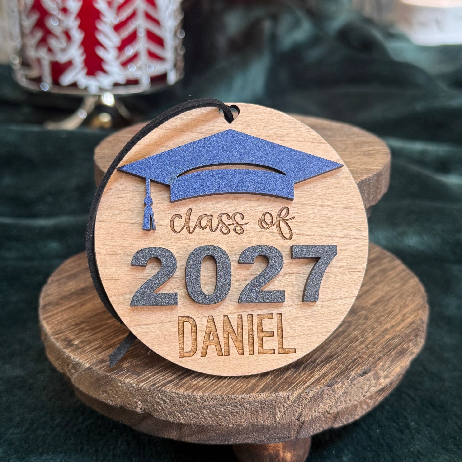 Class of 2027 wooden ornament in cherry finish with navy blue graduation cap, raised black 2027 numbers, and engraved name Daniel