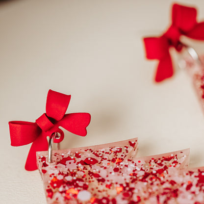 Close of red bows on Red and white glitter acrylic Christmas tree earrings with red bow studs, lightweight with 2 inch drop length, holiday dangles styled on a neutral background.