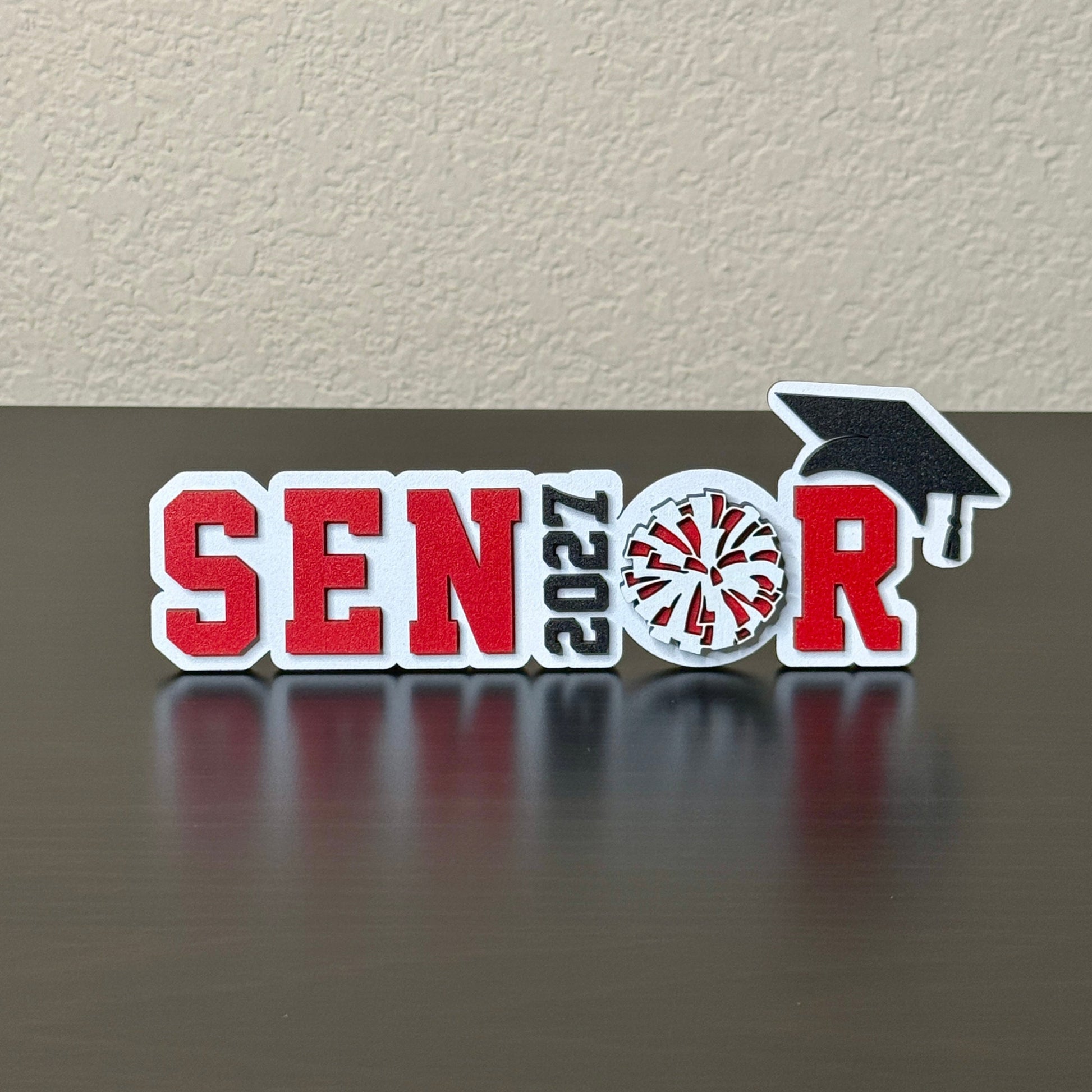Freestanding Class of 2027 graduation tabletop sign featuring SENIOR design with pom pom detail with a white wood base.