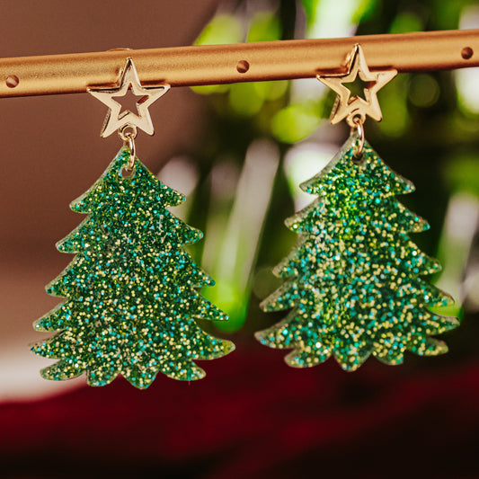 Green and gold glitter acrylic Christmas tree earrings with gold star studs hanging on display rod, showing sparkling festive dangle design by Half Turn Designs