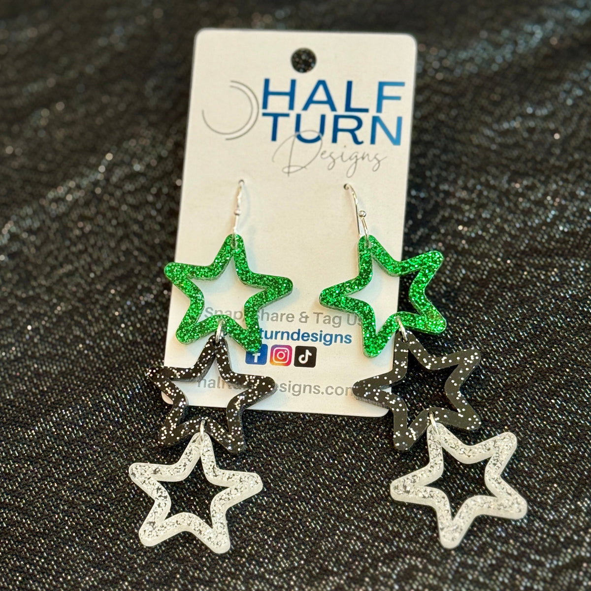 Stars dangle earrings made from three dangling stars of green glitter, black glitter and white glitter on silver earrings hooks by half turn designs.