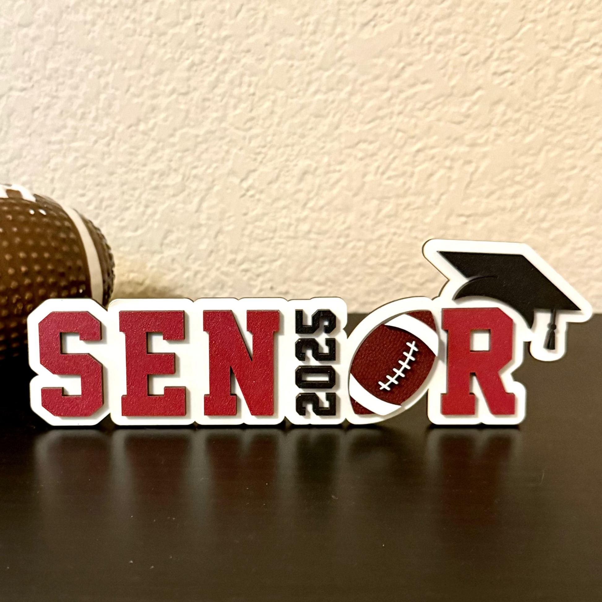 High School Senior football sign measuring 7" in length by 2.35 inches in height.   White wood background with black graduation cap and year or graduation numbers, brown and white football and customers choice of colors for the letters SENR, which is part of the wood senior, perfect for graduation parties and football banquets.