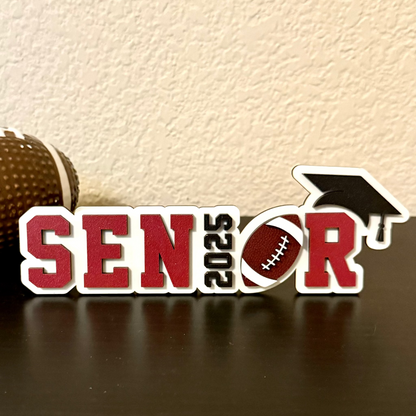High School Senior football sign measuring 7" in length by 2.35 inches in height.   White wood background with black graduation cap and year or graduation numbers, brown and white football and customers choice of colors for the letters SENR, which is part of the wood senior, perfect for graduation parties and football banquets.