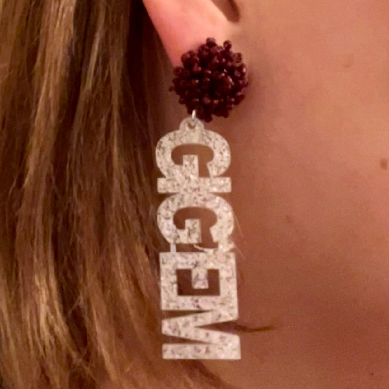 A model wearing officially licensed Texas A&M Dangle Earrings that spell out Gig 'em in white glitter with maroon seed bead toppers