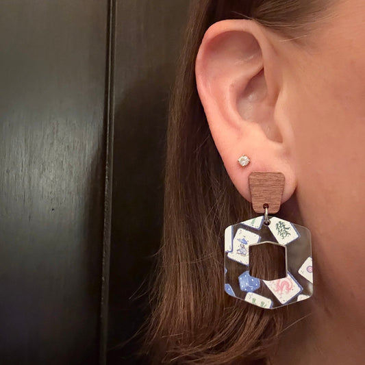 Model wearing Hexagon-shaped mahjong earrings made from lightweight clear acrylic printed with blue chinoiserie mahjong tiles and dice, featuring a hollow center and paired with angular walnut wood studs.