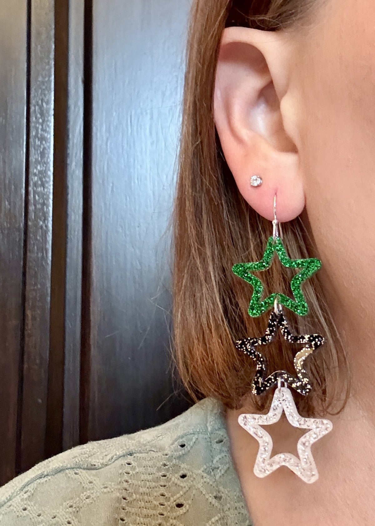 Model wearing Triple star dangle earrings featuring green glitter top stars, black speckled middle stars, and silver-glitter acrylic bottom stars on silver hooks