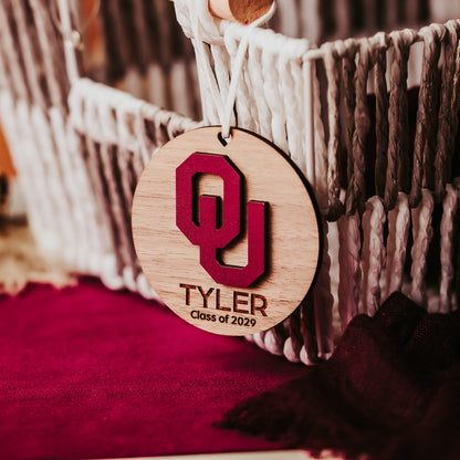 Engraved University of Oklahoma ornament featuring crimson OU logo on 3-inch round wood ornament, customizable with name or phrase, available in Cherry, Mahogany, Walnut, Sapele, or White finishes. Perfect Sooner Christmas gift or car charm. Shown here in walnut.;
