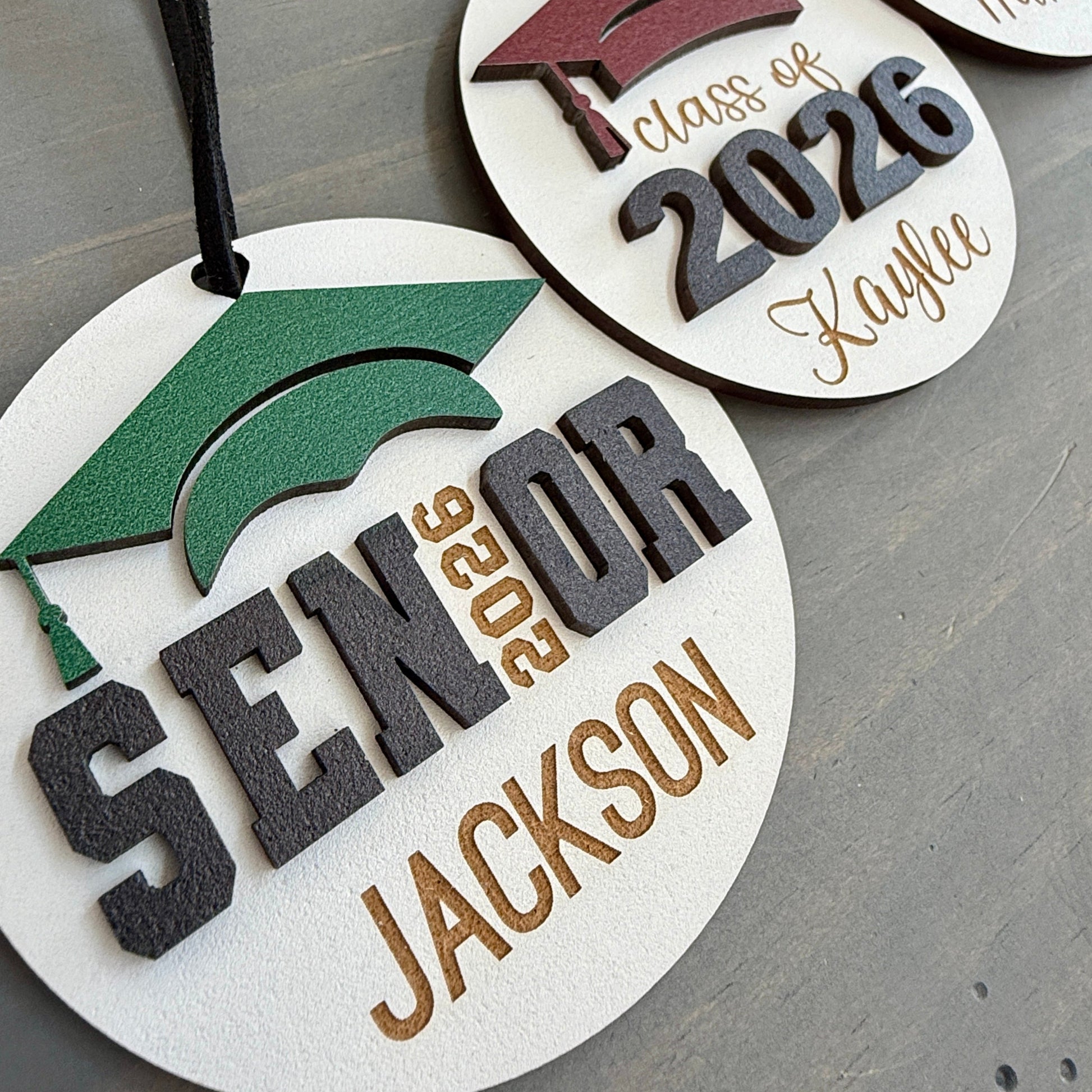 Personalized Class of 2026 ornament in white wood, engraved with the high school senior graduates name in three of the available styles from Half Turn Designs. 14 high school colors are available to further personalize the ornament with your choice of graduation cap color.