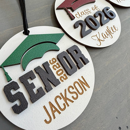 Personalized Class of 2026 ornament in white wood, engraved with the high school senior graduates name in three of the available styles from Half Turn Designs. 14 high school colors are available to further personalize the ornament with your choice of graduation cap color.