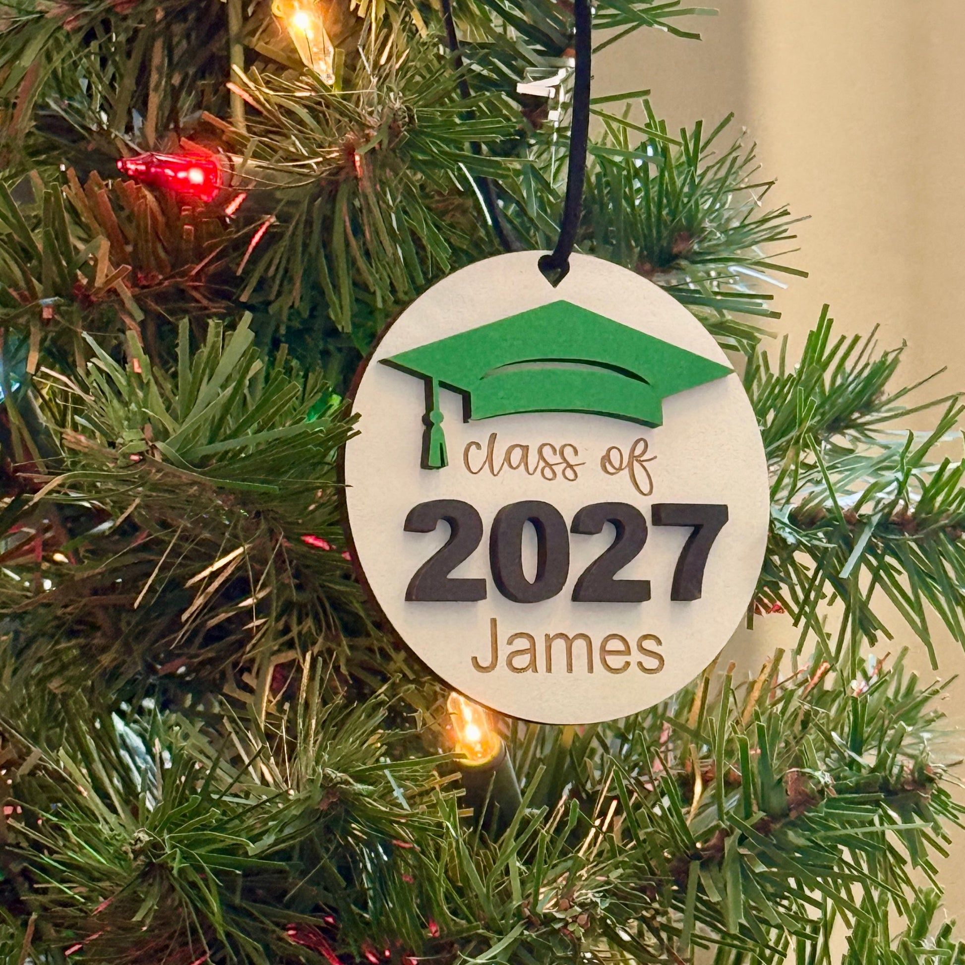 Personalized Class of 2027 wooden ornament in white finish with green graduation cap, raised black 2027 numbers and graduate's engraved name