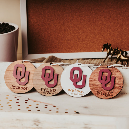 Engraved University of Oklahoma ornament featuring crimson OU logo on 3-inch round wood ornament, customizable with name or phrase, available in Cherry, Mahogany, Walnut, Sapele, or White finishes. Perfect Sooner Christmas gift or car charm. Shown here, from left to right in Cherry, Walnut, White & Mahogany