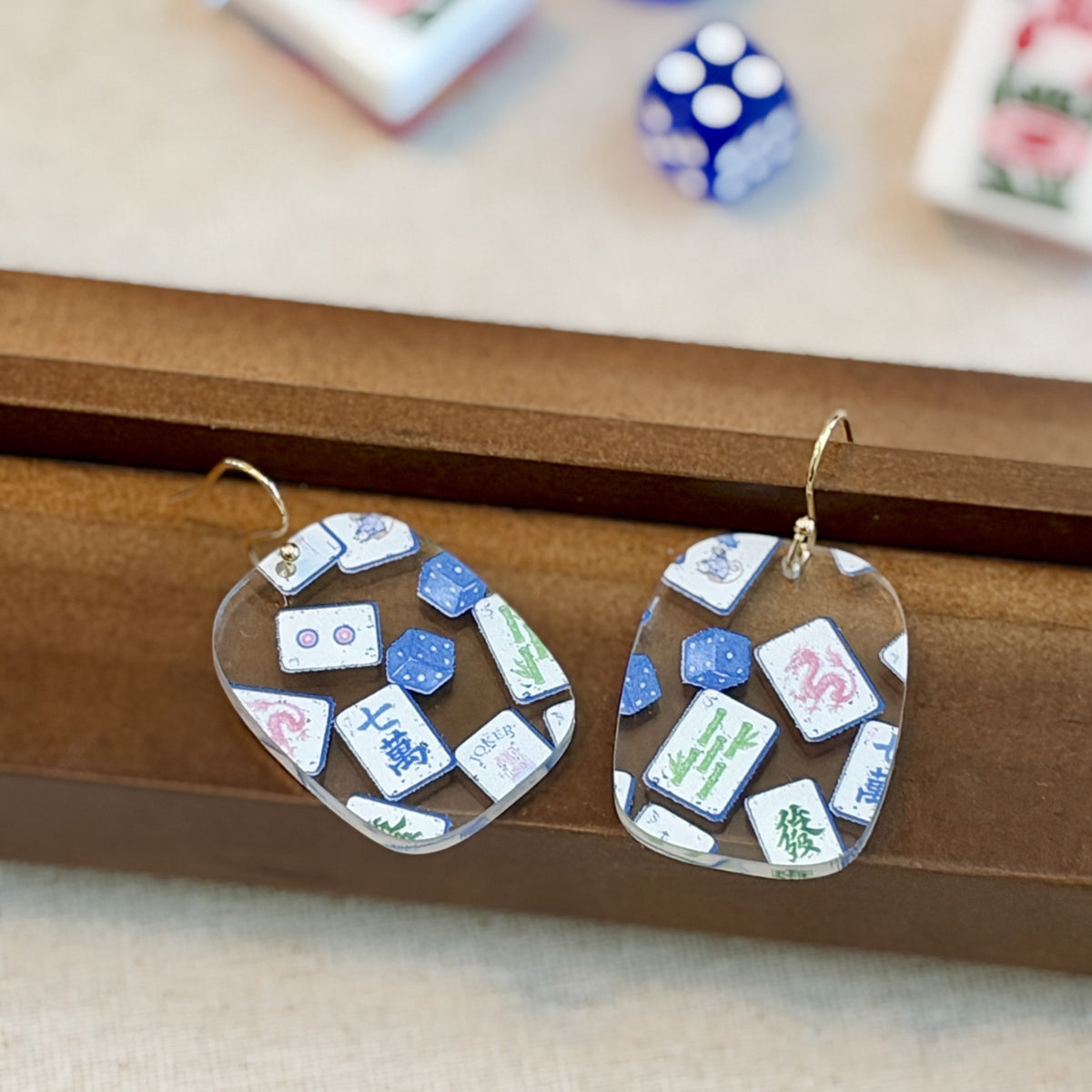 Blue chinoiserie mahjong tile earrings in clear acrylic with Jokers and dice on gold-tone hooks, beside mahjong tiles and racks