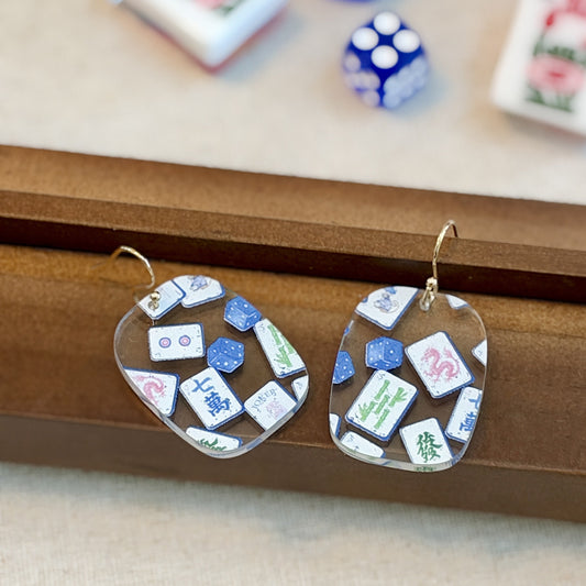 Blue chinoiserie mahjong tile earrings in clear acrylic with Jokers and dice on gold-tone hooks, beside mahjong tiles and racks