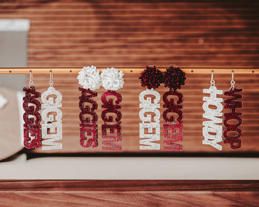 Texas A&M Officially Licensed Mix and Match Dangle Earrings in maroon and white glitter. Create your own pair of earrings combining TAMU phrases like "Aggies", "Gig 'em", "Whoop" and "Howdy" into a pair of earrings with your choice of glitter color and earring topper.