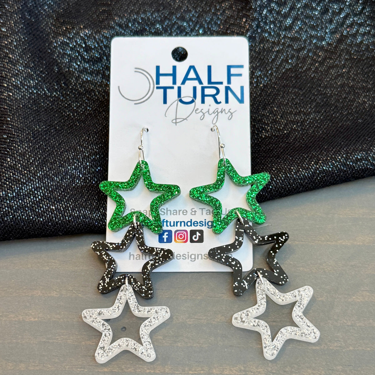 Triple star dangle earrings featuring green glitter top stars, black speckled middle stars, and silver-glitter acrylic bottom stars on silver hooks, displayed on a Half Turn Designs card.