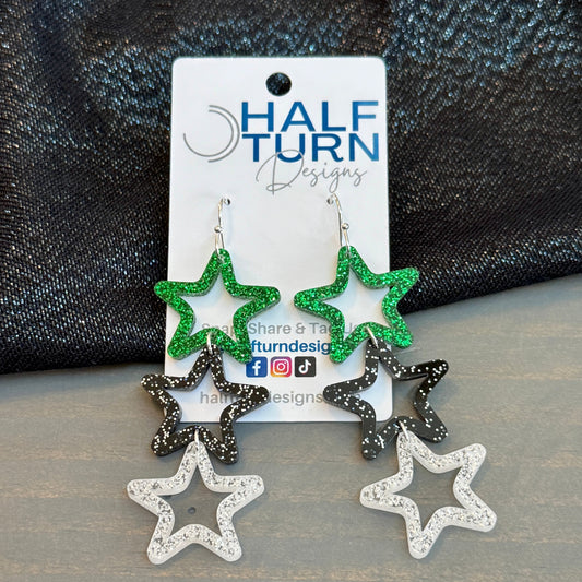 Triple star dangle earrings featuring green glitter top stars, black speckled middle stars, and silver-glitter acrylic bottom stars on silver hooks, displayed on a Half Turn Designs card.