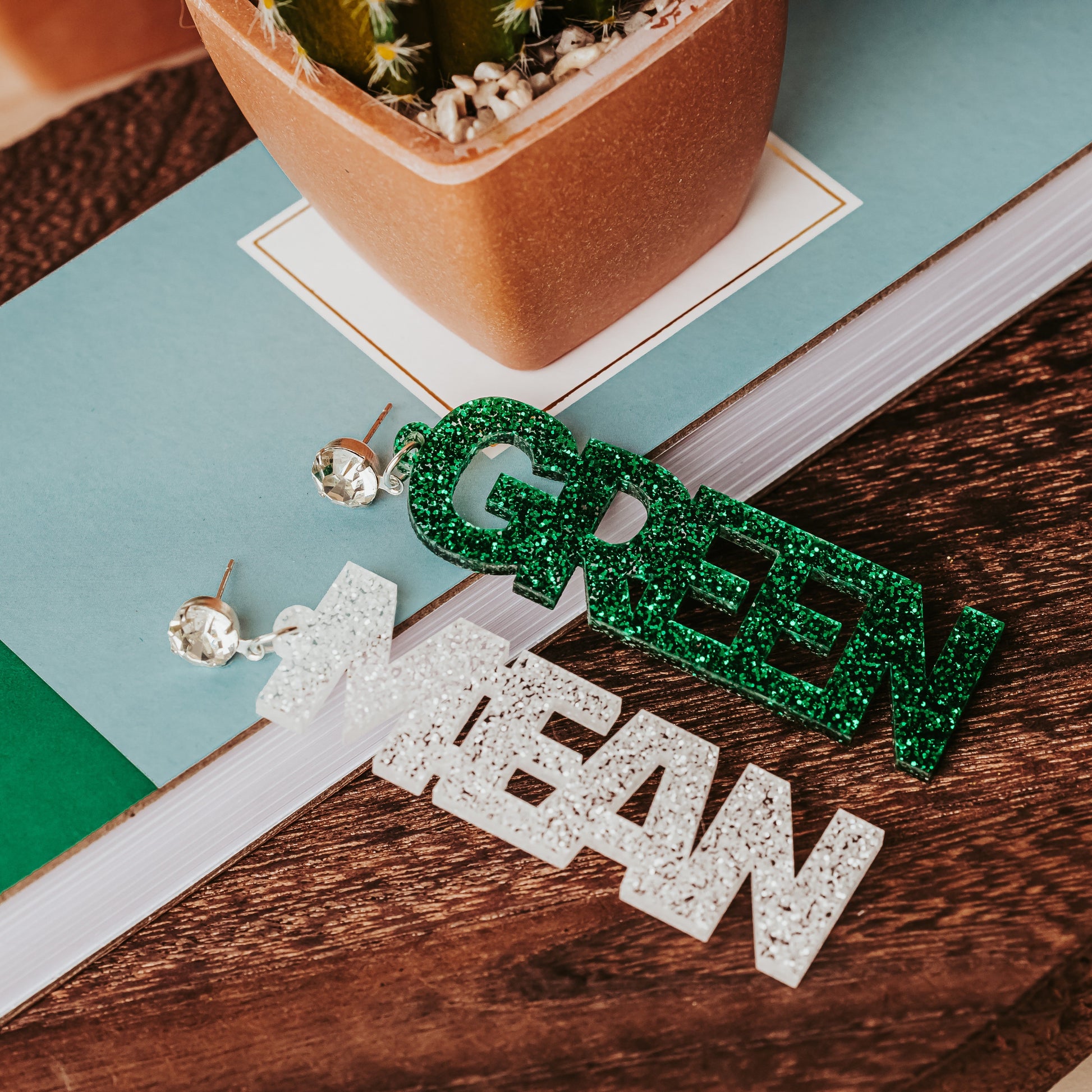 Officially licensed University of North Texas Mean Green Game Day earrings in green and white glitter with your choice of earrings topper including silver earring hooks, rhinestone topper, white tube bead topper, green tinsel topper, iridescent tinsel topper or white tinsel topper.  Mean and Green earrings shown here in green glitter with silver stainless steel earring hooks. Mean shown here in white glitter and green shown in green glitter with rhinestone earring toppers
