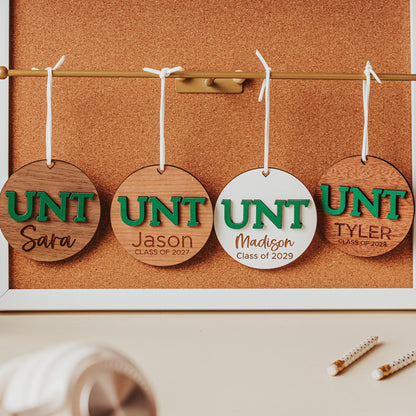 University of North Texas ornaments featuring green UNT letters with engraved personalization.  Shown in four wood types, from left to right, walnut, cherry, white and mahogany.  Officially licensed Mean Green personalized ornament by Half Turn Designs.